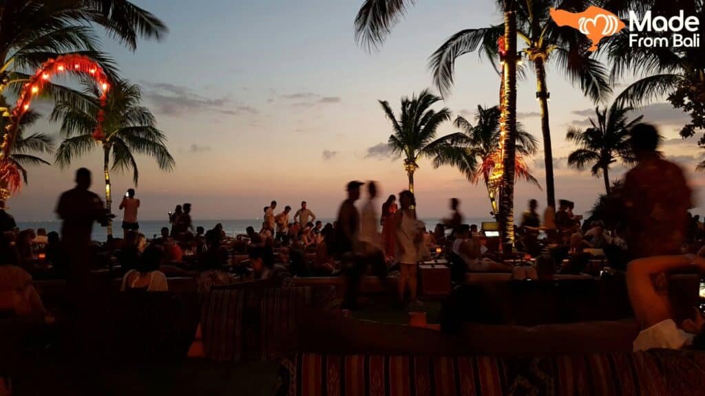 Crowded Bali beach club at sunset with palm trees, ocean view, and guests relaxing by the seaside.