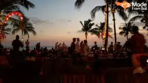 Crowded Bali beach club at sunset with palm trees, ocean view, and guests relaxing by the seaside.