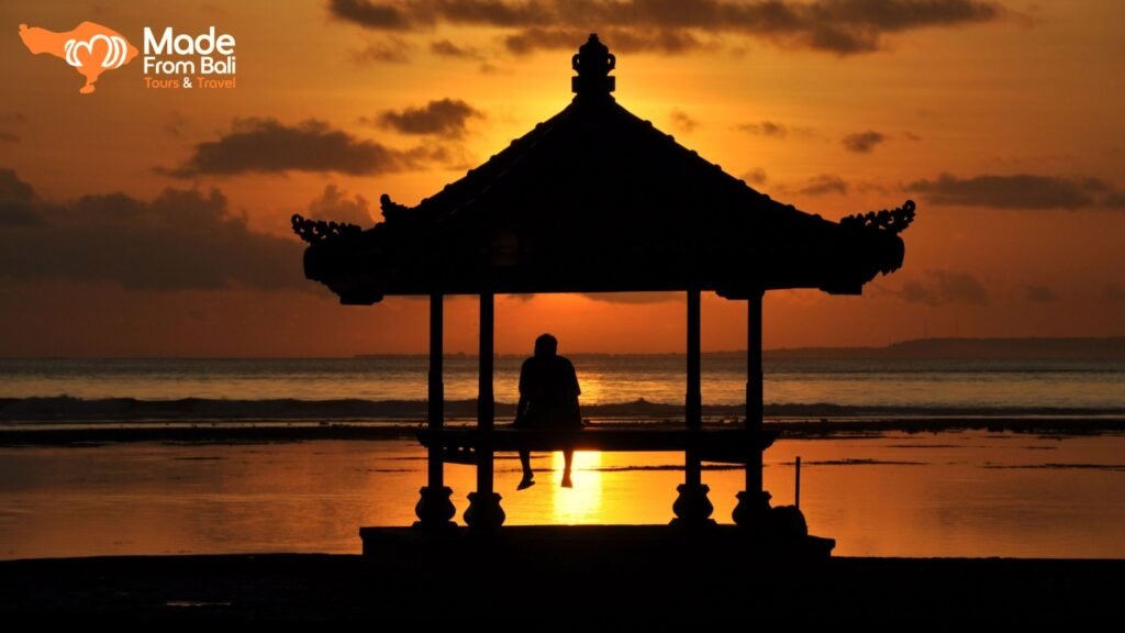 Person sitting in a beach pavilion, watching a colorful sunset over the ocean in Bali.