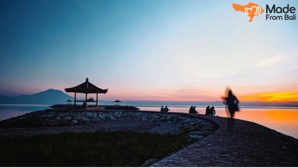 Gazebo tepi pantai Sanur saat matahari terbit, dengan pengunjung duduk menikmati suasana laut yang tenang.