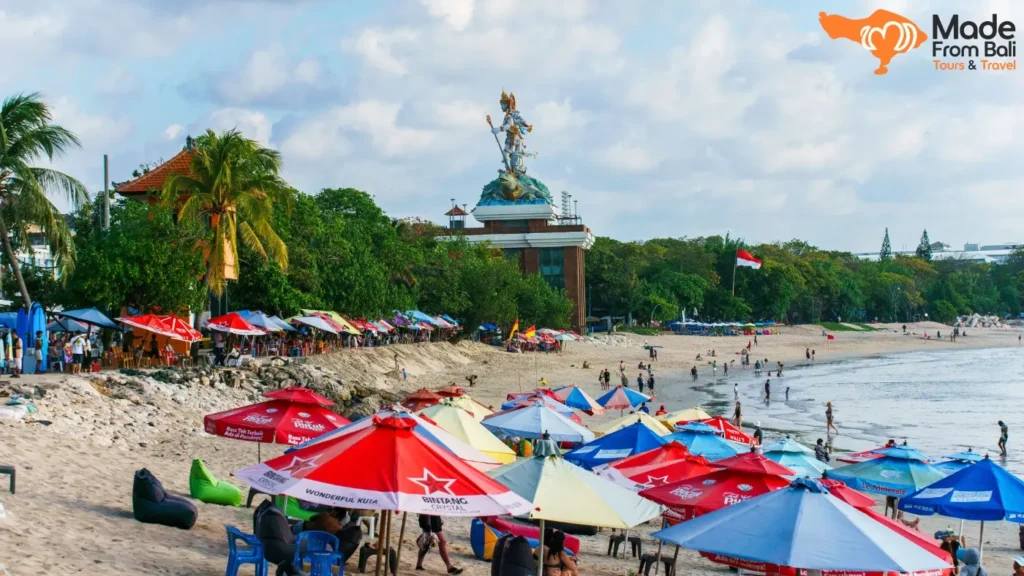 Suasana Pantai Kuta dengan payung warna-warni, wisatawan, dan garis pantai yang dekat menuju kawasan Legian.