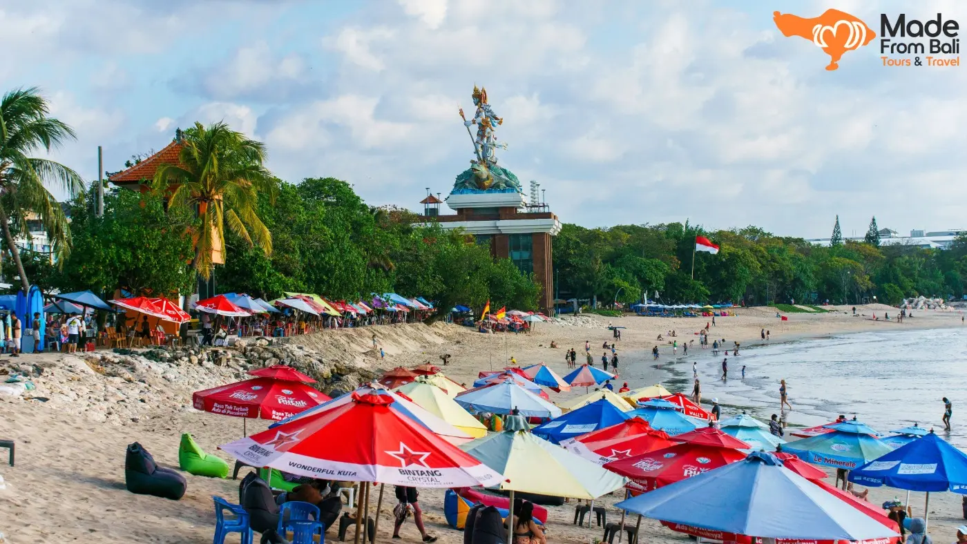 Suasana Pantai Kuta dengan payung warna-warni, wisatawan, dan garis pantai yang dekat menuju kawasan Legian.